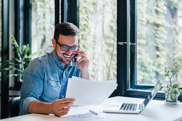 Portrait of young smiling cheerful entrepreneur in casual office making phone call while working with charts and graphs looking at paper documents